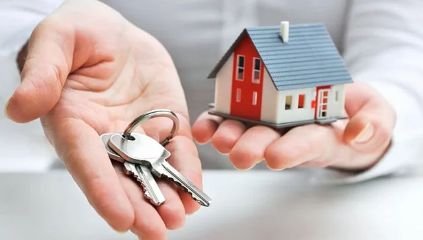 A person holds a set of silver house keys in one hand and a small model of a house with a red roof in the other, symbolizing homeownership.
