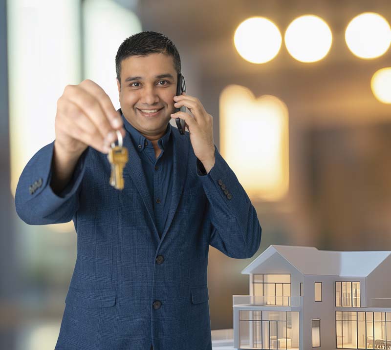A smiling man in a blue suit holds keys and a phone, standing beside a model house. The blurred background has warm, glowing lights, conveying optimism.