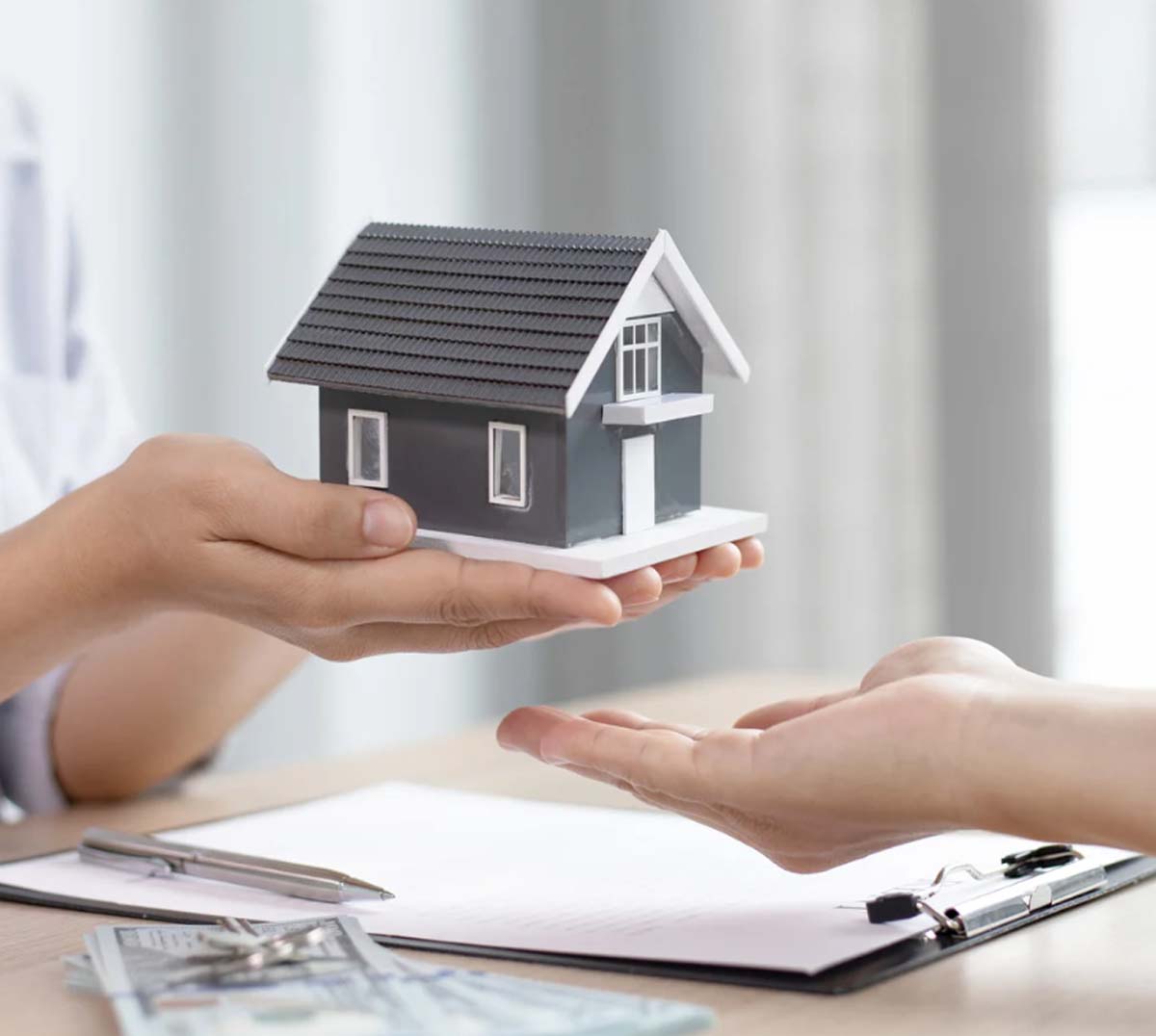 Hands exchanging a model house above a clipboard and money on a table, symbolizing a real estate transaction or home purchase agreement.