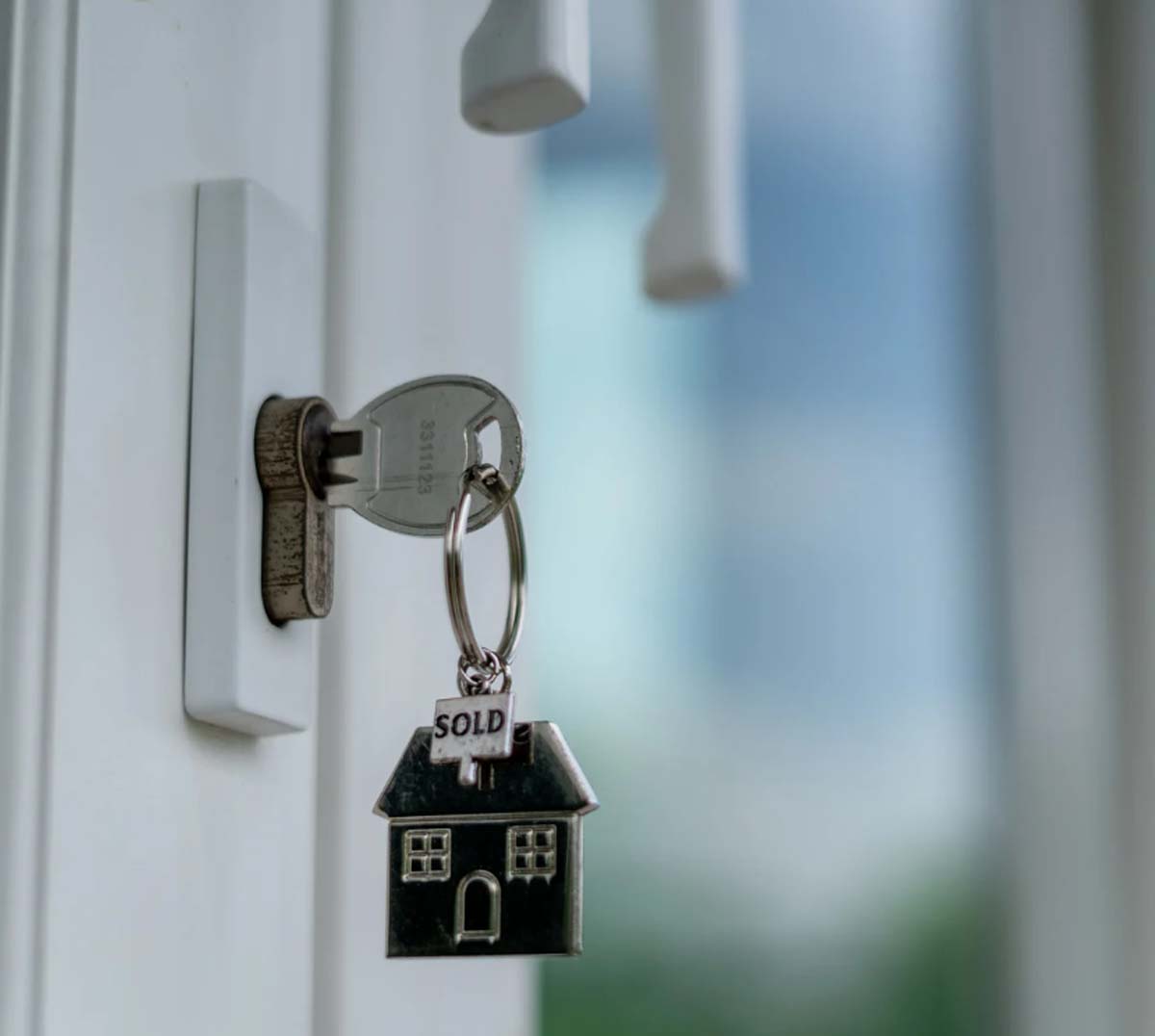 A key with a house-shaped keychain labeled "SOLD" is inserted in a door lock. The background is softly blurred, creating a sense of new beginnings.