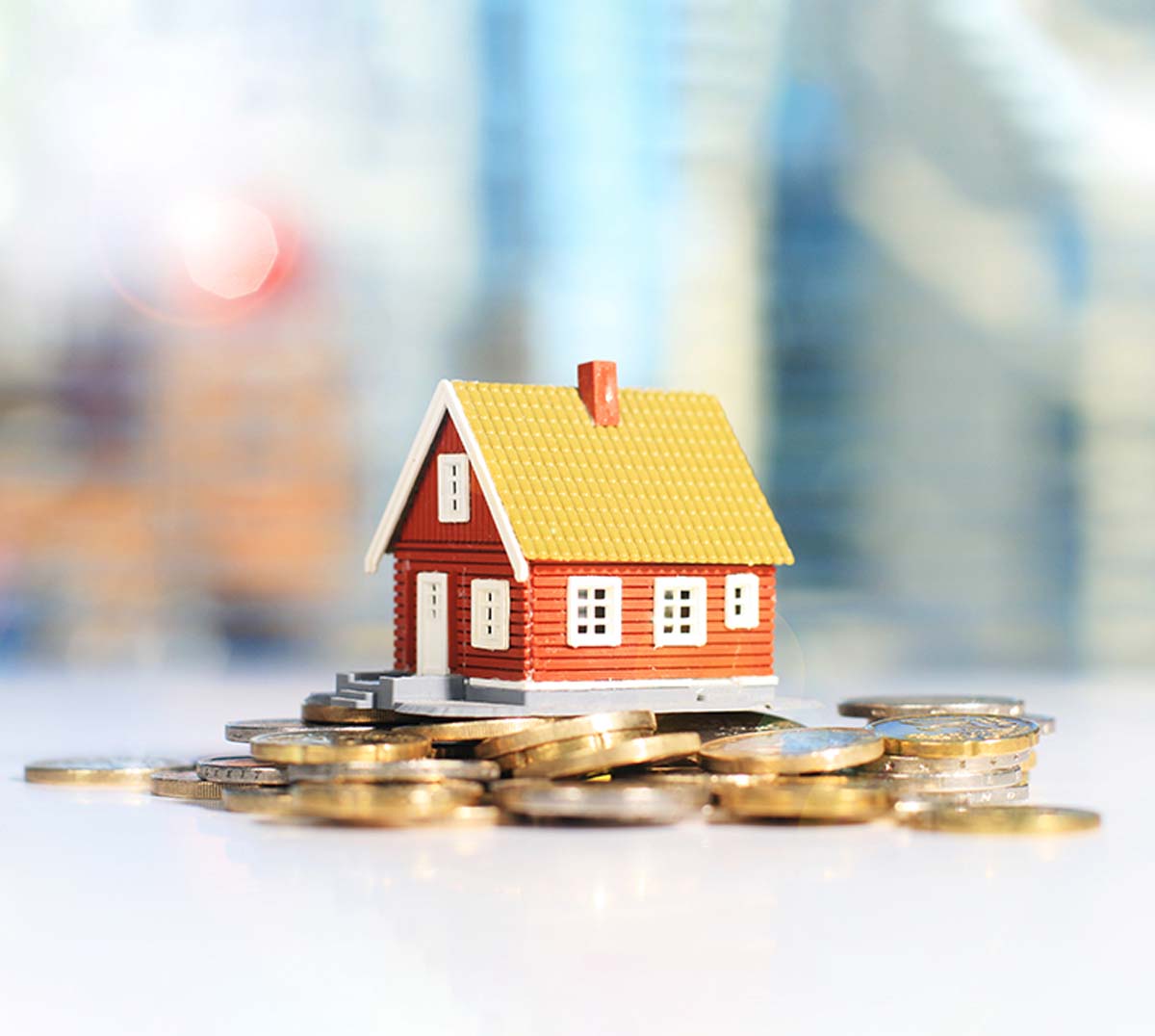 A small red house model with a yellow roof sits atop a pile of coins, symbolizing real estate investment. The blurred background adds a sense of depth.